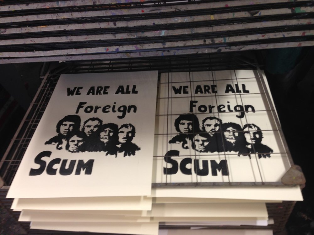 Screen printed white posters on a drying rack with the words "We are all foreign scum" around five people's faces