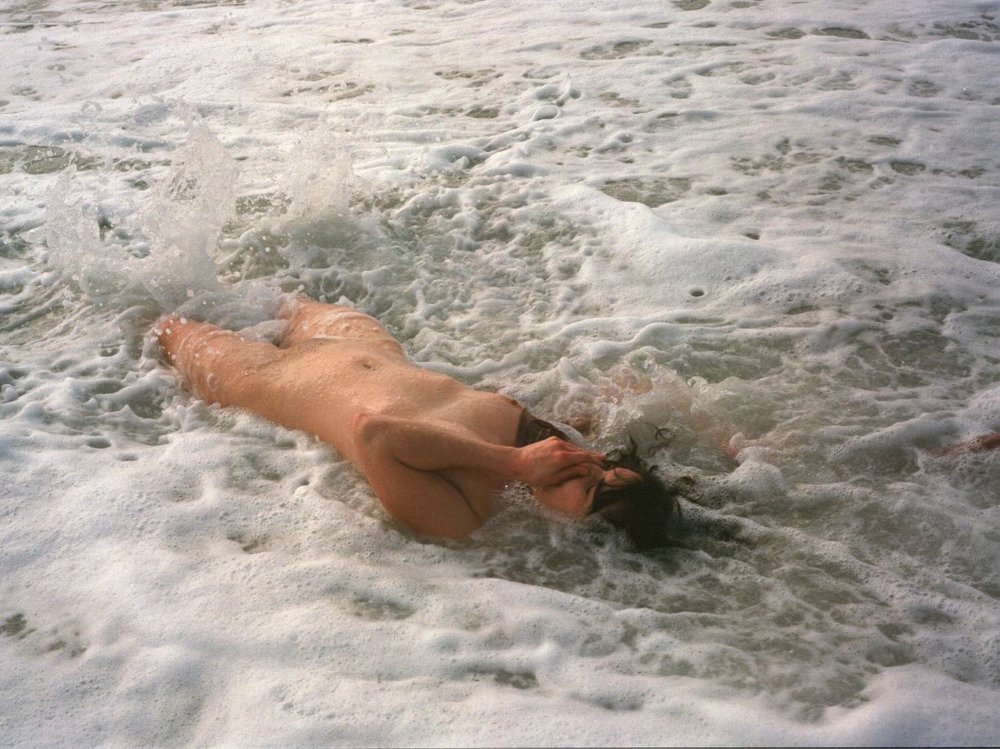 A woman lying on an ocean beach naked holding her nose as the waves crash over her